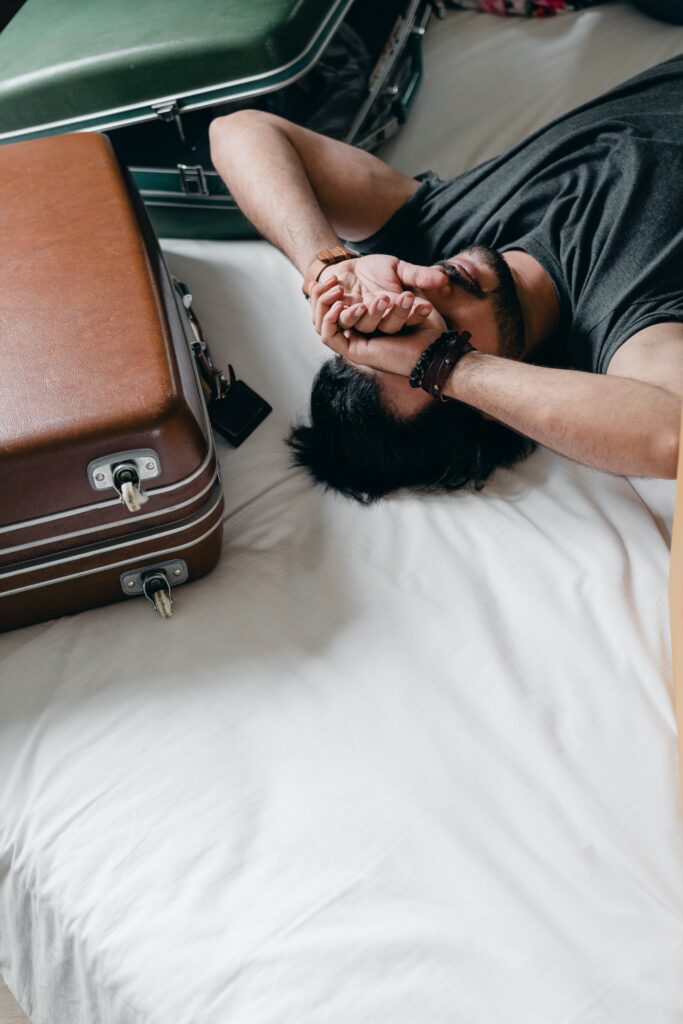 A man lying on a white bed with suitcases around him. He has his hands over his face like he is exasperated at all the paperwork to get married in the UK